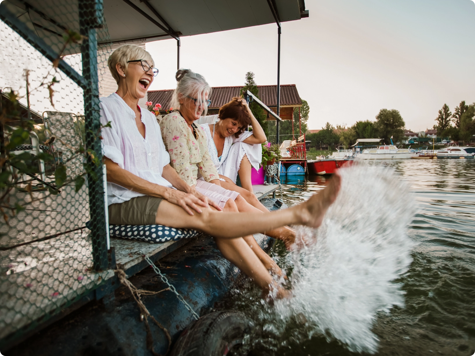 Three adult women sitting on a boat within a harbor with their legs hanging off the boat side kicking the water playfully.