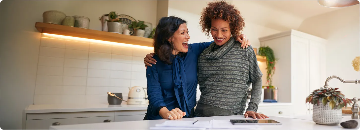 Two women standing together and laughing in a kitchen, suggesting midlife wellness and quality of life.